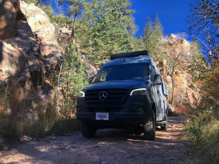 An off-road rv rental van on a dirt path, surrounded by rugged rocks and tall pine trees under a clear blue sky.. Winnebago Revel 2023