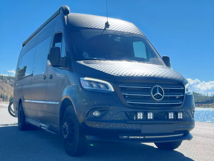 Dark Mercedes-Benz Sprinter Airstream RV rental van with geometric wrap, parked by a scenic lake under clear skies.. Airstream Interstate 2021