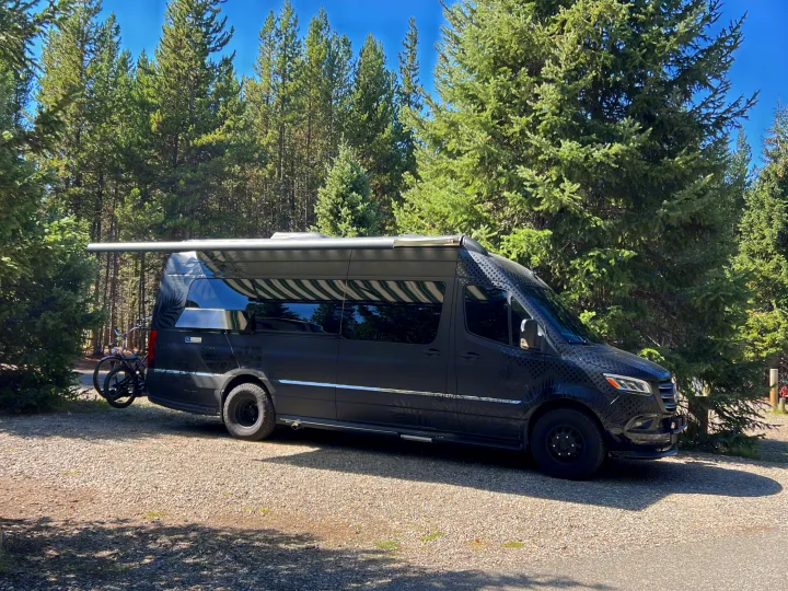 Dark grey Sprinter RV rental with awning extended, bike on back rack, parked on gravel in a sunny pine forest.. Airstream Interstate 2021