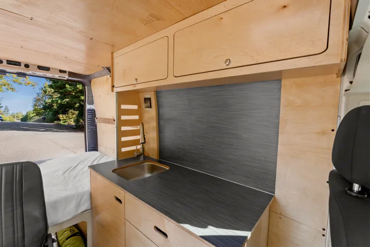 Inside a spacious RV rental: wood-paneled interior features a well-designed kitchen with sink, cabinetry, and comfortable bed.. RAM ProMaster 2022