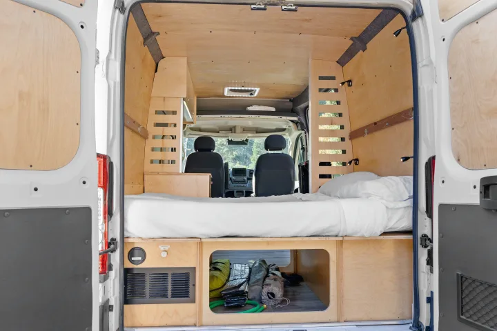 RV rental interior view from rear: a custom camper van conversion featuring a cozy bed, wood paneling, and storage.. RAM ProMaster 2022