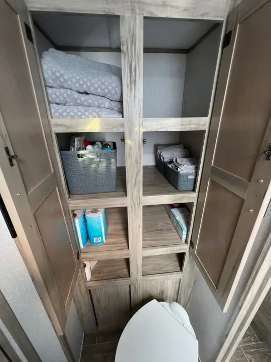 RV rental bathroom storage cabinet with open doors, showing neatly organized shelves with blankets, toiletries, & toilet paper.. Forest River Cherokee Alpha Wolf 2021