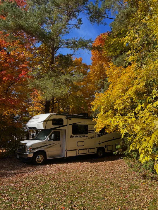 A Sunseeker RV rental is parked in a vibrant autumn forest, with red, orange, and yellow leaves under a blue sky.. Forest River Sunseeker 2023