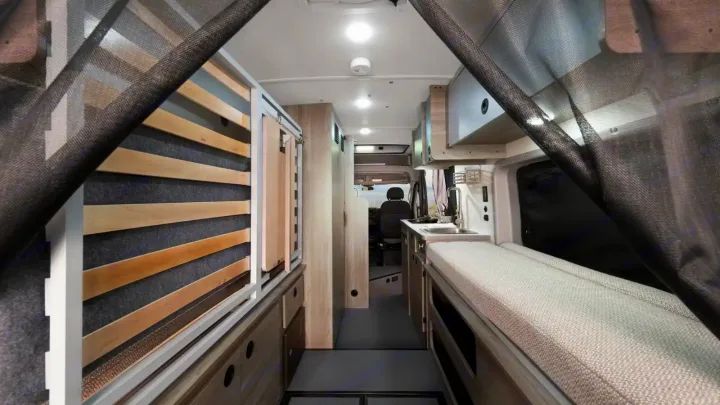 Interior of an RV rental showing a narrow aisle, convertible bed, kitchen, and black mesh privacy screens.. Winnebago Solis 2021
