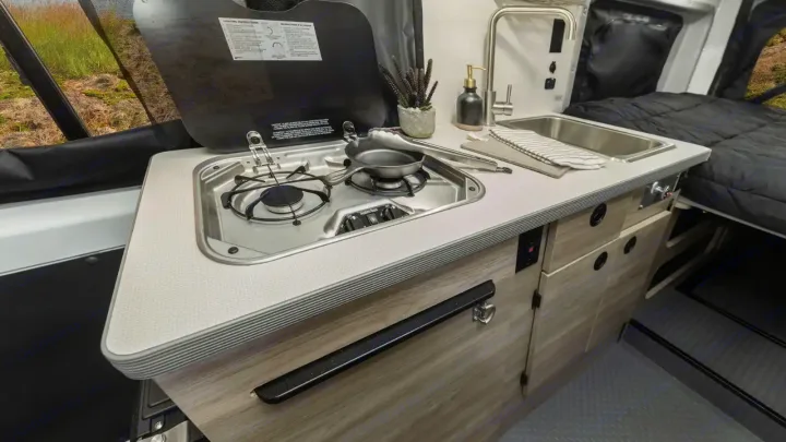 This RV rental offers a well-equipped compact kitchen with a stove, sink, and storage, adjacent to a comfortable bed.. Winnebago Solis 2021