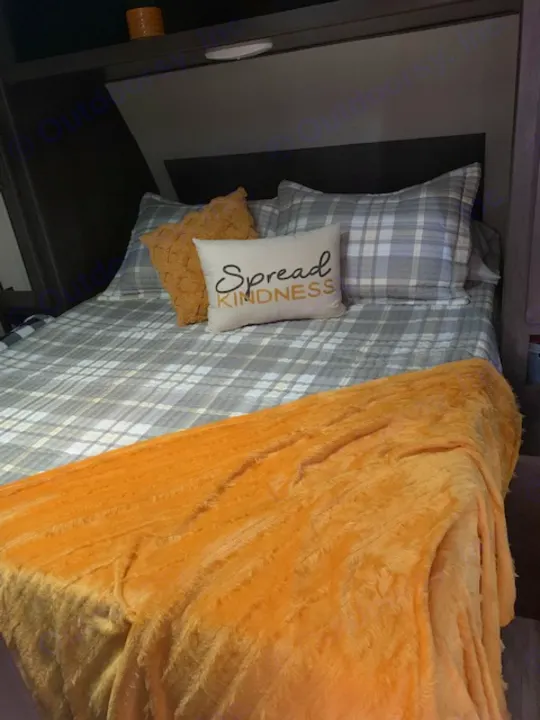 Inside an RV rental: a cozy bedroom features plaid bedding, a fluffy orange blanket, and a "Spread Kindness" pillow.. Grand Design Transcend Xplor 2022
