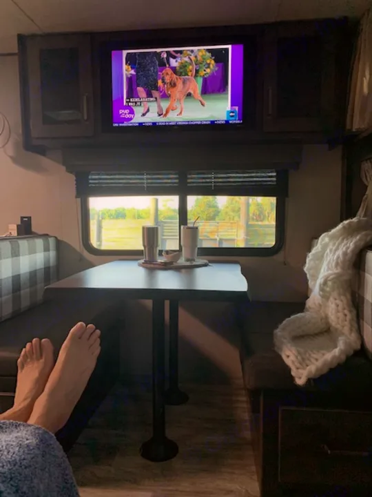Enjoying a cozy RV rental: feet up, watching a dog show on TV, with a table and window view inside.. Grand Design Transcend Xplor 2022