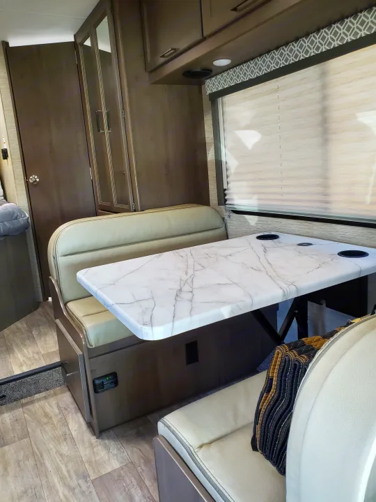 RV rental dinette: Light-toned interior with a marble-look table, upholstered benches, window blinds, and wood-look flooring.. Thor Coleman 2023