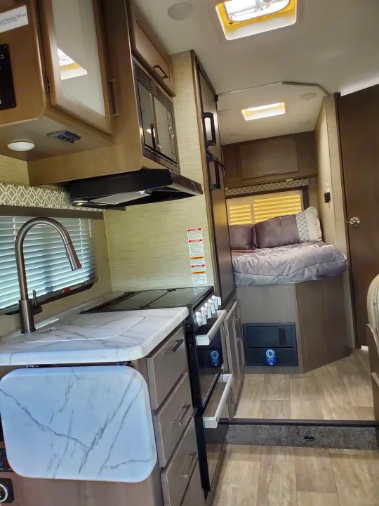 This RV rental interior features a compact kitchen with sink, stove, microwave, fridge, and an elevated bed area.. Thor Coleman 2023