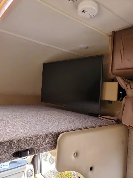 An RV rental's over-cab bunk area with a mounted Insignia TV, showing safety features like a smoke detector.. Thor Coleman 2023