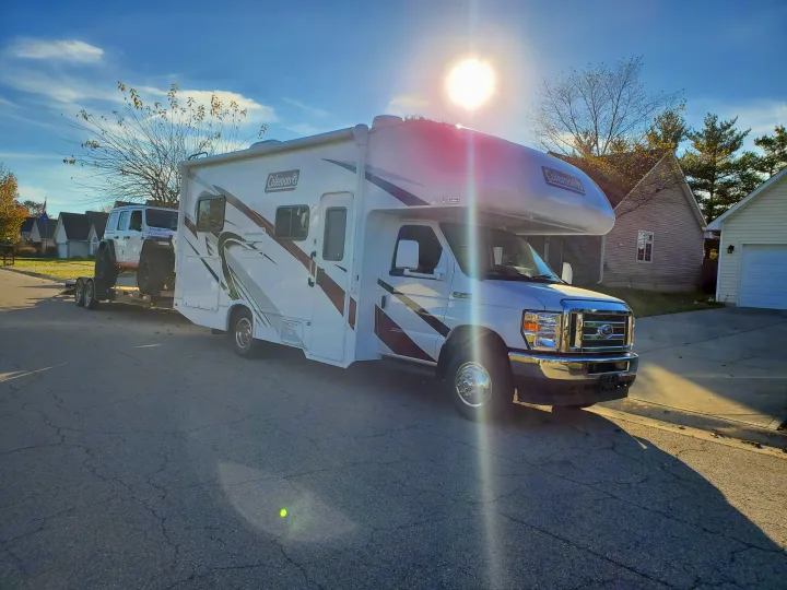 White Coleman RV rental towing a white Jeep on a trailer down a sunny residential street.. Thor Coleman 2023