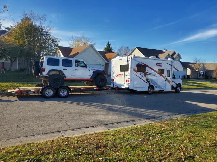 White RV rental (Coleman) tows a white Jeep Wrangler on a flatbed trailer down a suburban street.. Thor Coleman 2023