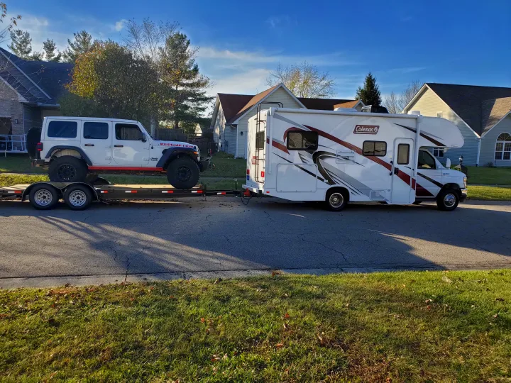 A white Coleman RV rental is towing a white Jeep Wrangler on a flatbed trailer.. Thor Coleman 2023
