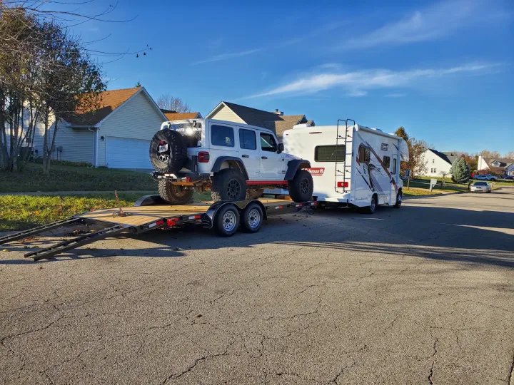 White Jeep on trailer, towed by an RV rental. Ready for adventure from a sunny residential street with birds in sky.. Thor Coleman 2023