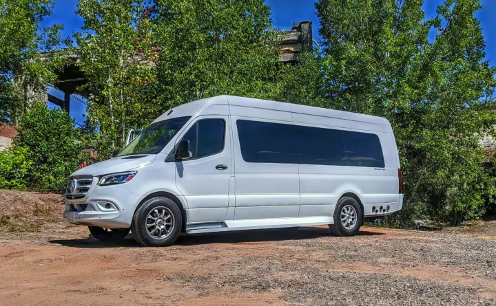 This sleek white Mercedes Sprinter van, ideal for RV rental, is parked on a gravel lot with trees and old structures behind it.. Ultimate Toys Coach- Midwest Automotive Daycruiser D6 2022