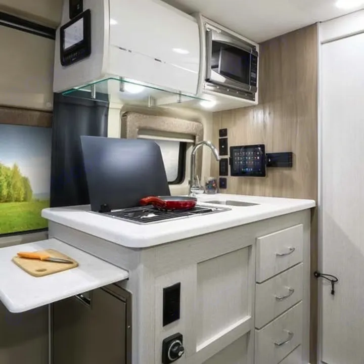 Functional rv rental kitchen: light wood, white counters, built-in stove, sink, microwave, tablet mount, and pull-out prep space.. Thor Rize 2022