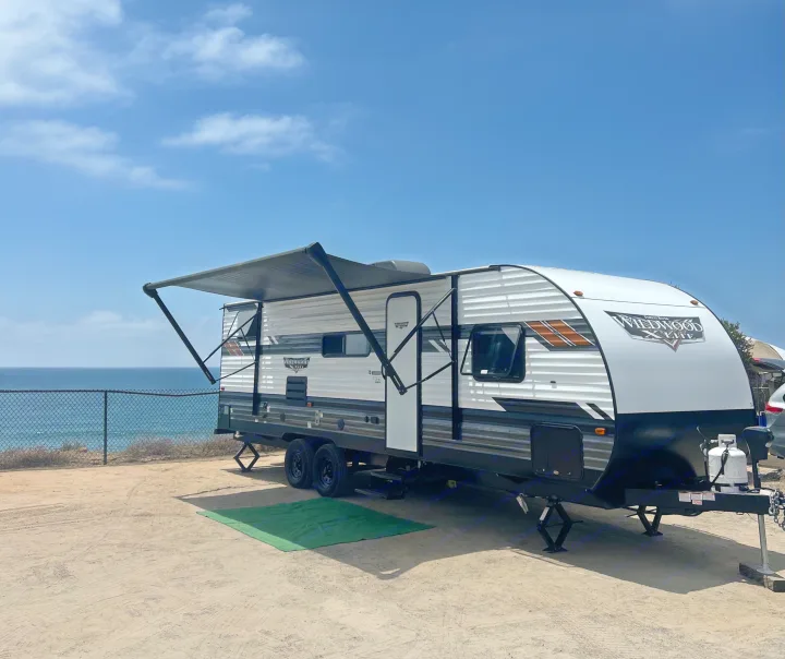 White RV travel trailer rental with awning extended, parked on a dirt lot overlooking the ocean under a clear blue sky.. Forest River Wildwood X-Lite 2023