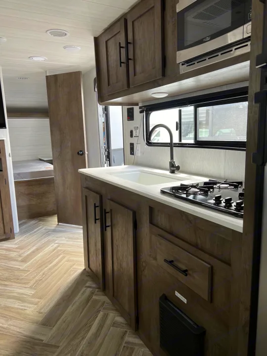 Modern RV rental kitchen with dark wood cabinets, stove, sink, oven, and light herringbone wood floors. Ideal for trips!. Forest River Wildwood X-Lite 2023