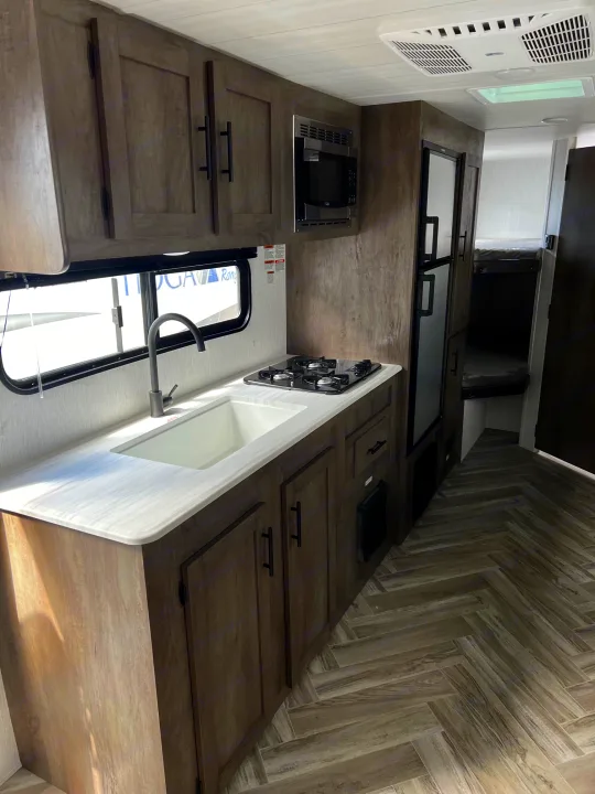 Modern RV rental kitchen showcasing microwave, stove, sink, and stylish wood-grain cabinets with herringbone flooring.. Forest River Wildwood X-Lite 2023