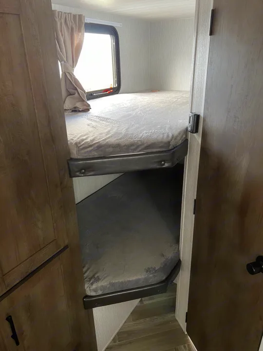 Looking up into an **rv rental's** sleeping area with two stacked bunk beds, featuring gray mattresses and a bright window with a curtain.. Forest River Wildwood X-Lite 2023