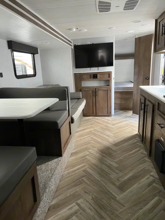 Inside an RV rental: view of a sleek living area with gray sofa, wall TV, wood-look herringbone floor, and light walls.. Forest River Wildwood X-Lite 2023