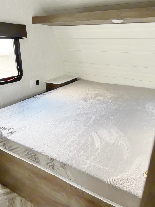 This cozy rv rental interior features a sleeping area with a silver comforter, light wood panels, & outlets.. Forest River Wildwood X-Lite 2023
