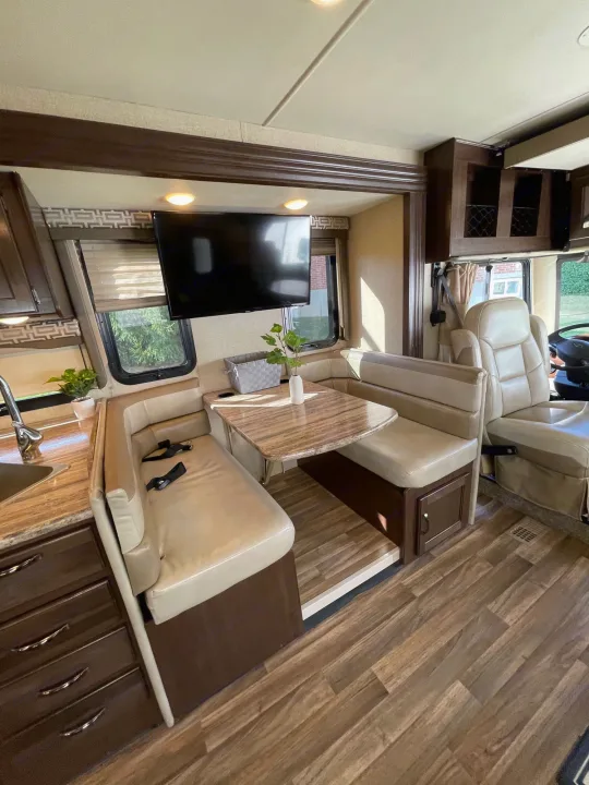 Bright RV rental interior with a light-toned dinette, kitchen counter, mounted TV, and front cab seating.. Thor A.C.E. 2017