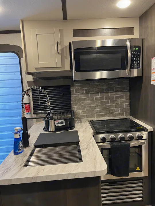 Inside a compact, modern RV rental kitchen, featuring a stainless steel microwave, stove, and sink with a sleek tiled backsplash.. Crossroads RV Cruiser Aire 2022