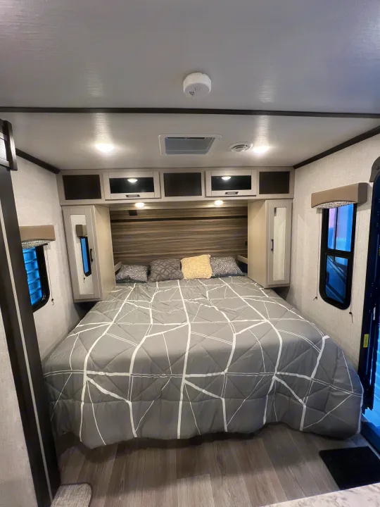 A modern RV rental bedroom with a grey geometric comforter on a large bed, overhead storage, and windows.. Crossroads RV Cruiser Aire 2022