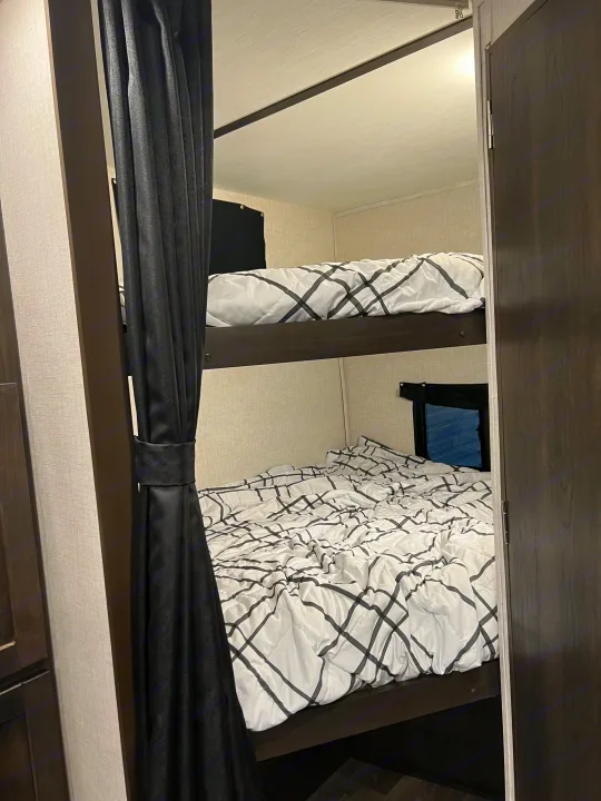 Two stacked bunk beds in an RV rental, with white patterned bedding, dark wood frames, and black curtains. Small windows are visible.. Crossroads RV Cruiser Aire 2022