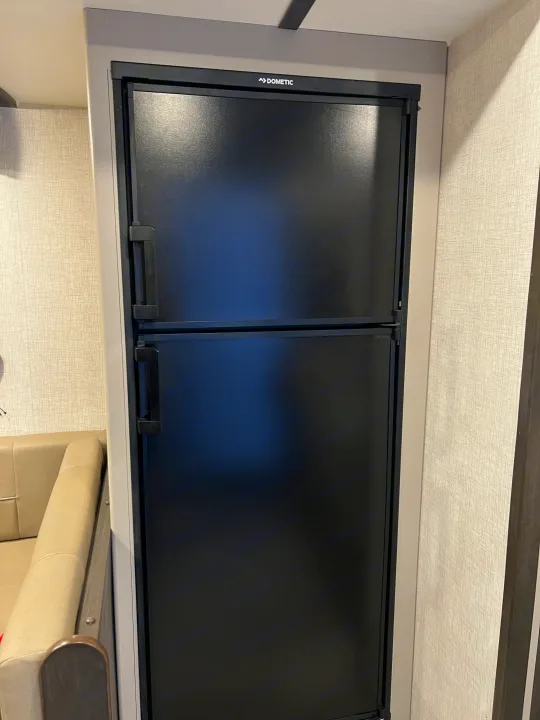 Black Dometic refrigerator in an RV rental. Top freezer, bottom fridge, built into the wall next to a beige sofa.. Crossroads RV Cruiser Aire 2022