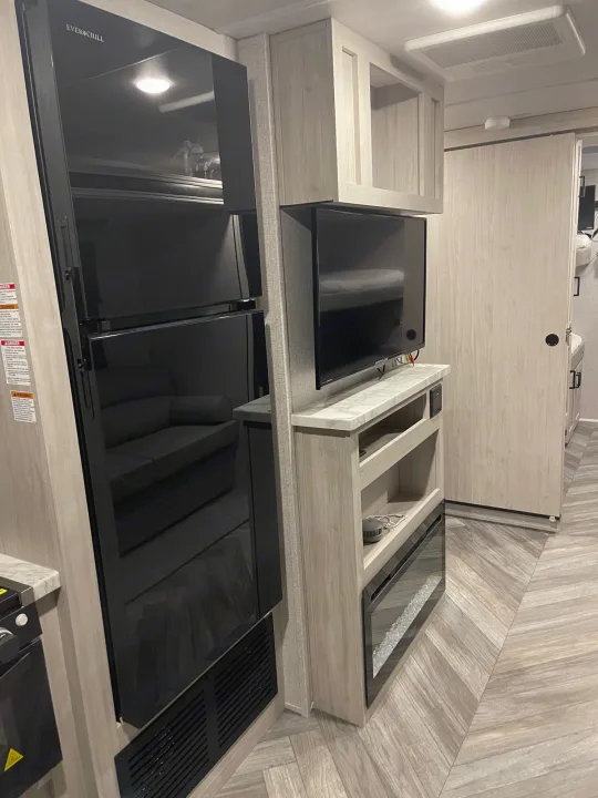 Modern RV rental interior: large black fridge, TV, light wood cabinetry, electric fireplace, and herringbone floor.. Della Terra 312H Della Terra 2023
