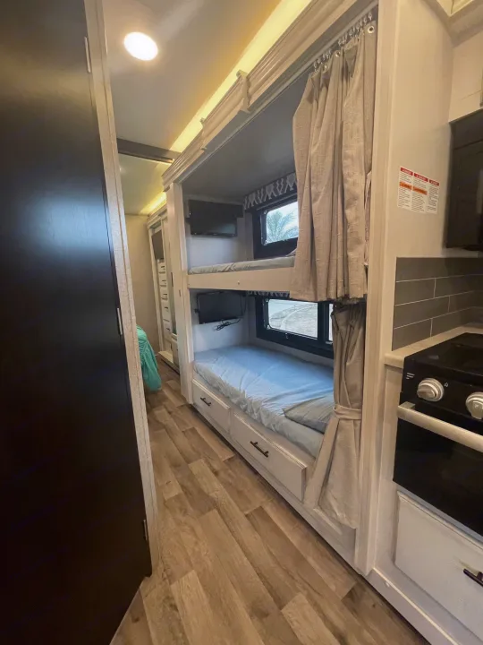 Inside an RV rental: Compact bunk beds with TVs & privacy curtains. A small kitchen with stove is visible right.. Entegra Coach Vision 29f Bunk house model 2021