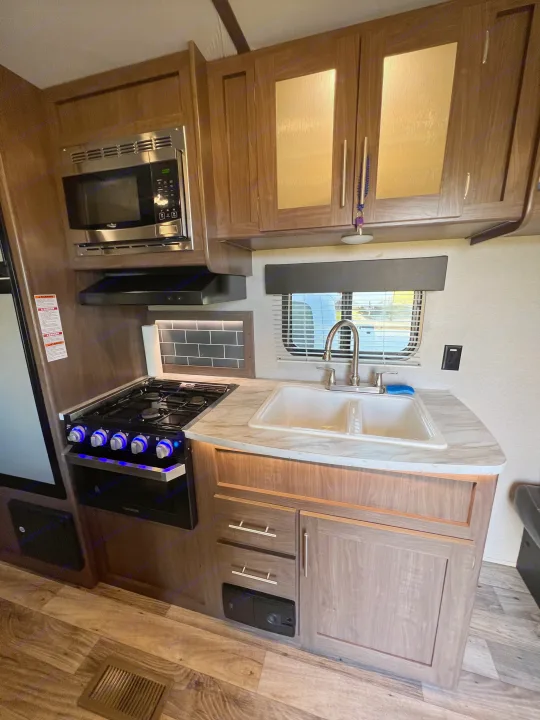A modern RV rental kitchen featuring a lit stove, microwave, double sink, and wood-look cabinets. Cook with ease on your trip!. Keystone RV Hideout LHS 2020