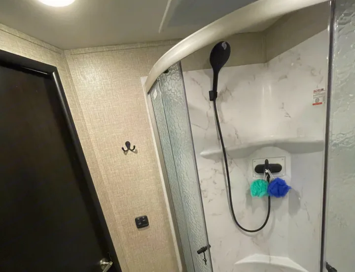 Inside an RV rental: a compact bathroom with a white marble-patterned shower, black showerhead, door, and wall hooks.. Entegra Coach Odyssey 2023