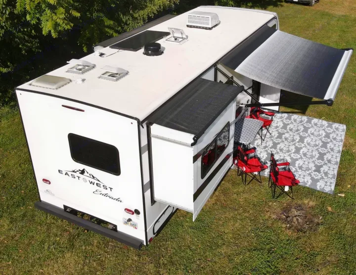This white **rv rental** is fully set up for camping, featuring an extended awning, outdoor rug, and chairs for relaxation.. East to West Entrada 2600DS 2022