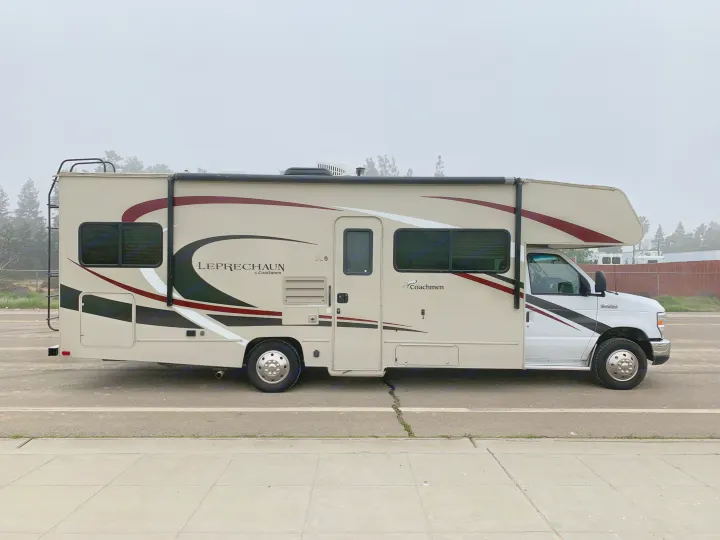 A light-colored Coachmen Leprechaun Class C motorhome, an ideal RV rental, with dark accent stripes, parked street-side.. Forest River Coachmen Leprechaun 2020