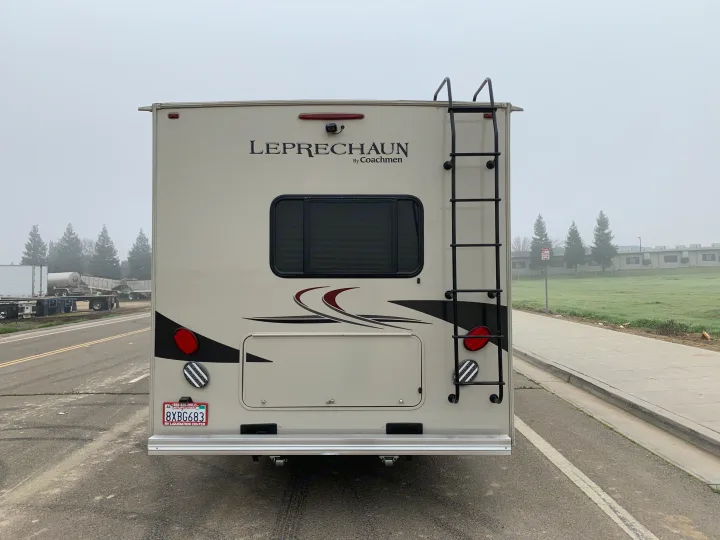 Rear of a beige Coachmen Leprechaun RV rental motorhome. Features a ladder, camera, and window, parked on a misty day.. Forest River Coachmen Leprechaun 2020