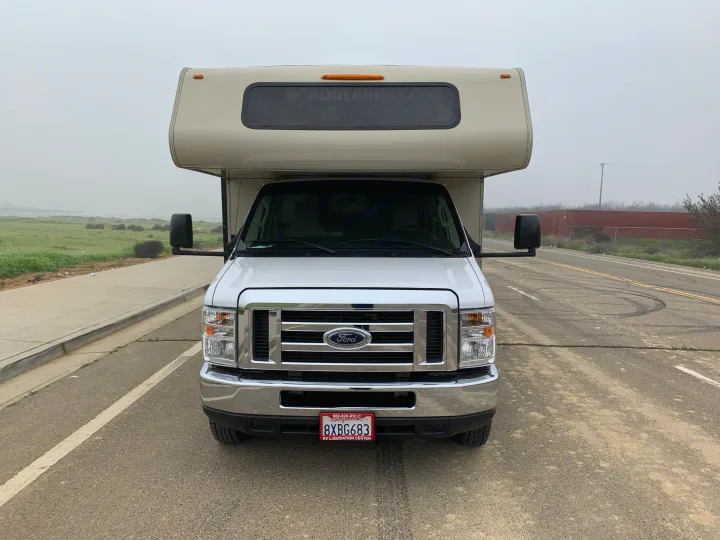 White Ford Class C RV rental facing forward, parked on an asphalt road. Ready for your next travel adventure in misty weather.. Forest River Coachmen Leprechaun 2020
