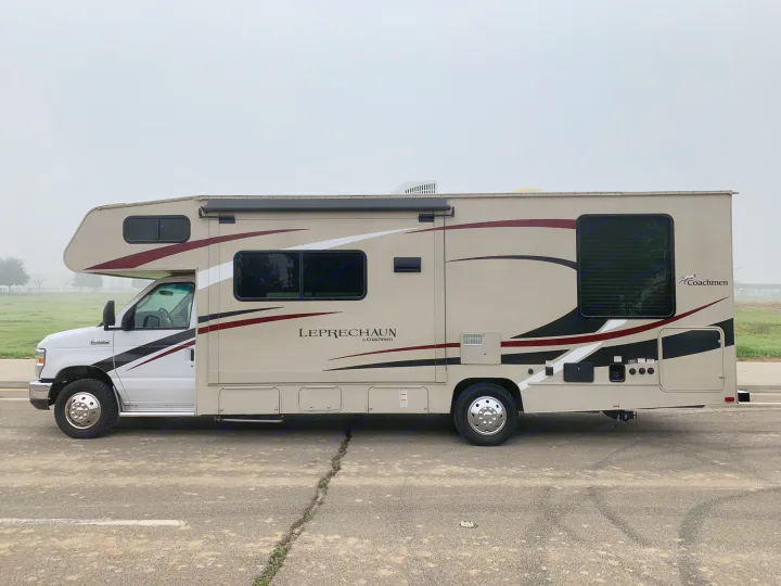 Side view of a large beige Coachmen Leprechaun RV rental, with stripe decals, parked on a cracked asphalt lot on a misty day.. Forest River Coachmen Leprechaun 2020