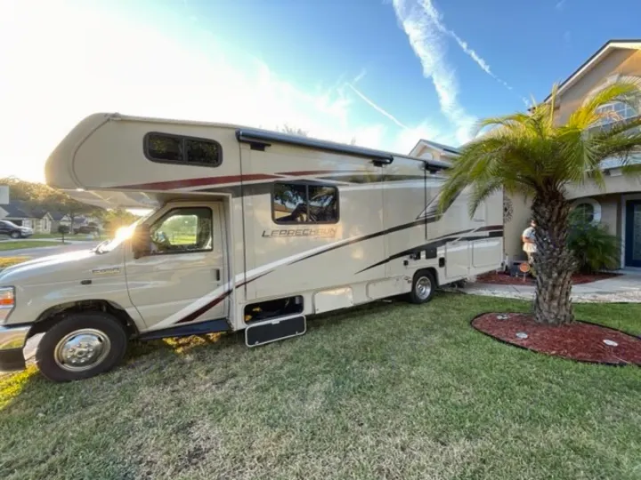 A beige Class C motorhome, an ideal RV rental, parked on a sunny lawn next to a house with an extended awning.. COACHMEN LEPRECHAUN 2021