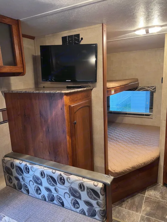 RV rental interior features a TV on a wood cabinet, a patterned sofa, and bunk beds with a window.. Keystone RV Hideout 2015