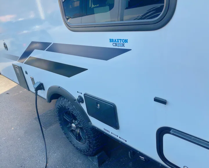 Side of a white Braxton Creek RV rental, connected to power. Shows tire and utility access for easy camping adventures.. Braxton Creek Bushwhacker Plus 2022