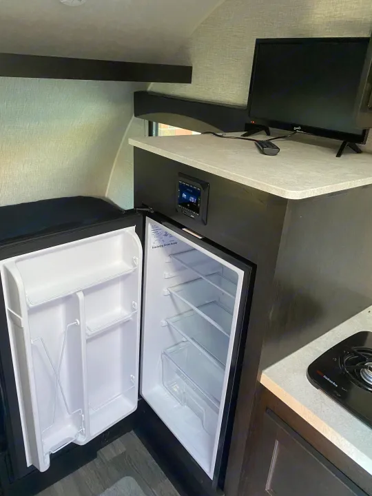 This **RV rental** interior features an open compact fridge, TV on counter, integrated stereo, & a cooktop. Ready for travel.. Braxton Creek Bushwhacker Plus 2022