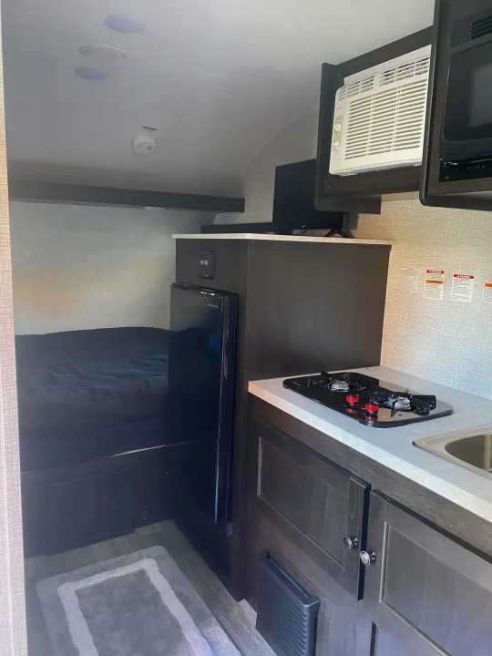 RV rental interior: Compact kitchen with stove, sink, and fridge; AC unit, microwave, TV, and cozy bed visible.. Braxton Creek Bushwhacker Plus 2022