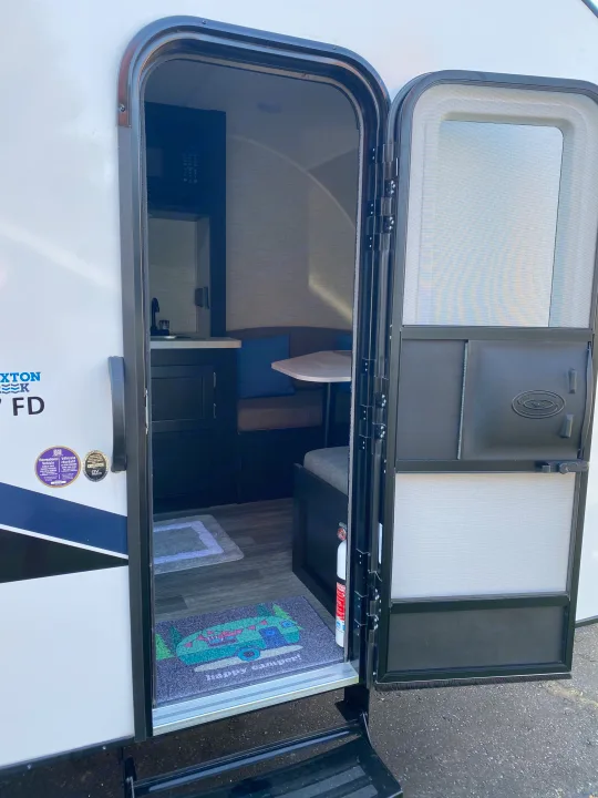 Explore this RV rental's interior. The open door reveals a compact kitchen, dining area, and a "happy camper" mat.. Braxton Creek Bushwhacker Plus 2022