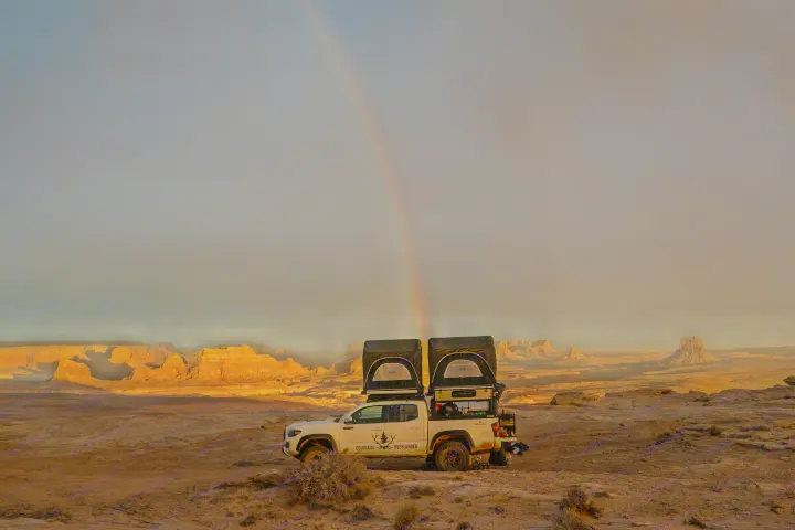 A white Toyota Tacoma, outfitted with rooftop tents like an RV rental, sits under a vibrant rainbow in a golden desert.. Toyota Tacoma 2022