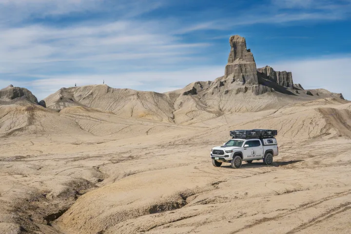 An overland truck, like an RV rental, explores a vast desert badlands landscape with towering rock formations & distant hikers.. Toyota Tacoma 2022