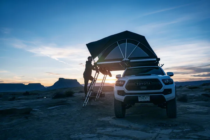 Rugged RV rental adventure: a Toyota Tacoma with a rooftop tent in a stunning desert sunrise. A person climbs into the elevated tent.. Toyota Tacoma 2022