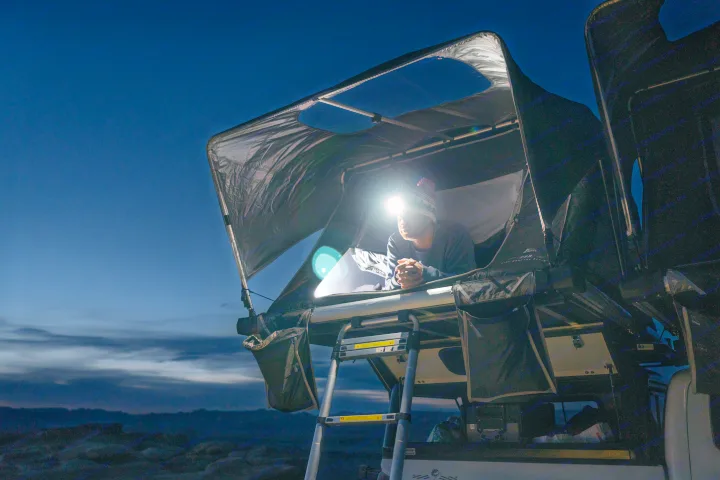 A man with a headlamp relaxes in a bright rooftop tent on an RV rental, gazing into the quiet dark night.. Toyota Tacoma 2022